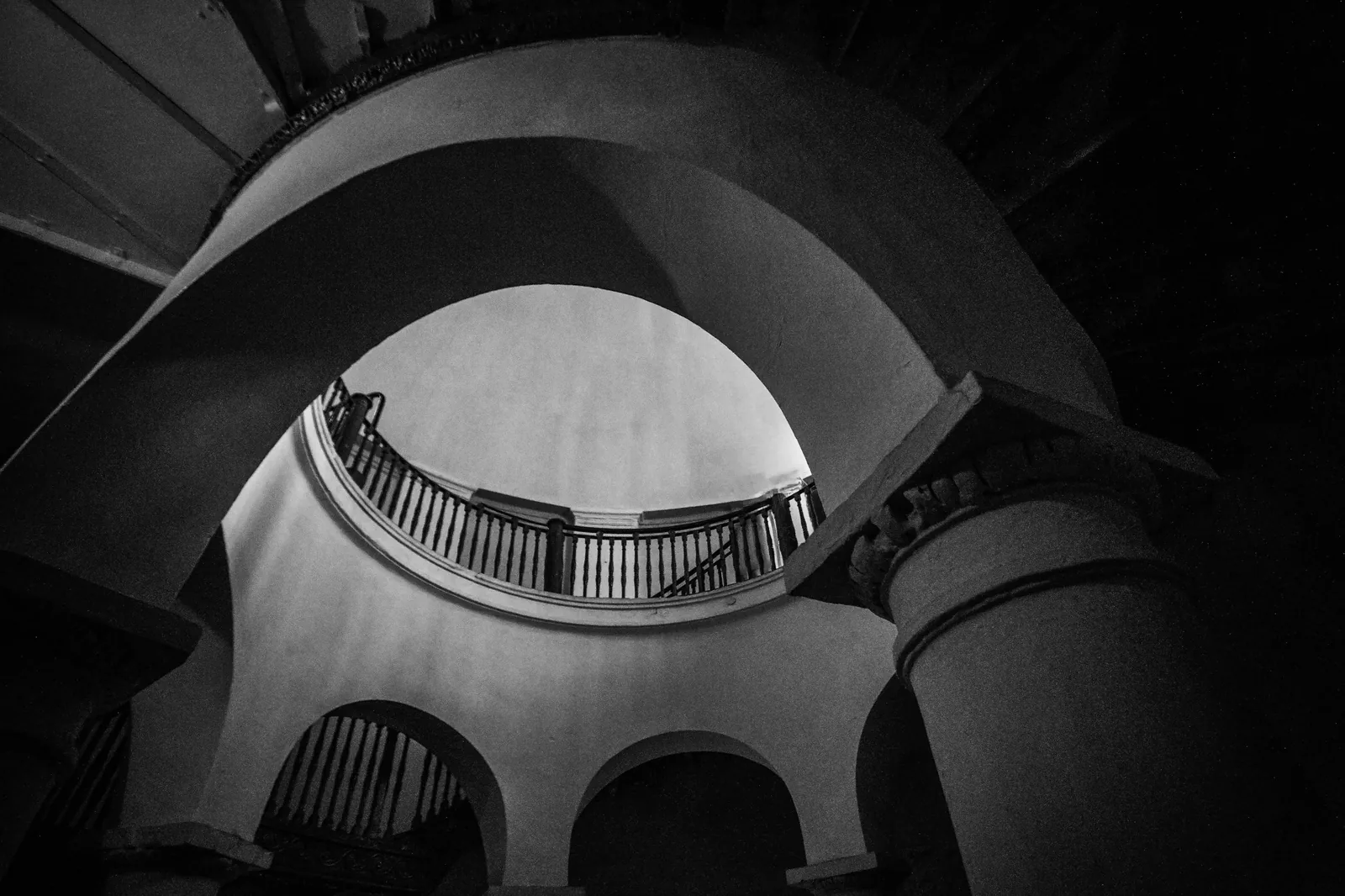 A grand spiral staircase photographed from below in black and white, its balustrade curving towards the light of the floor above