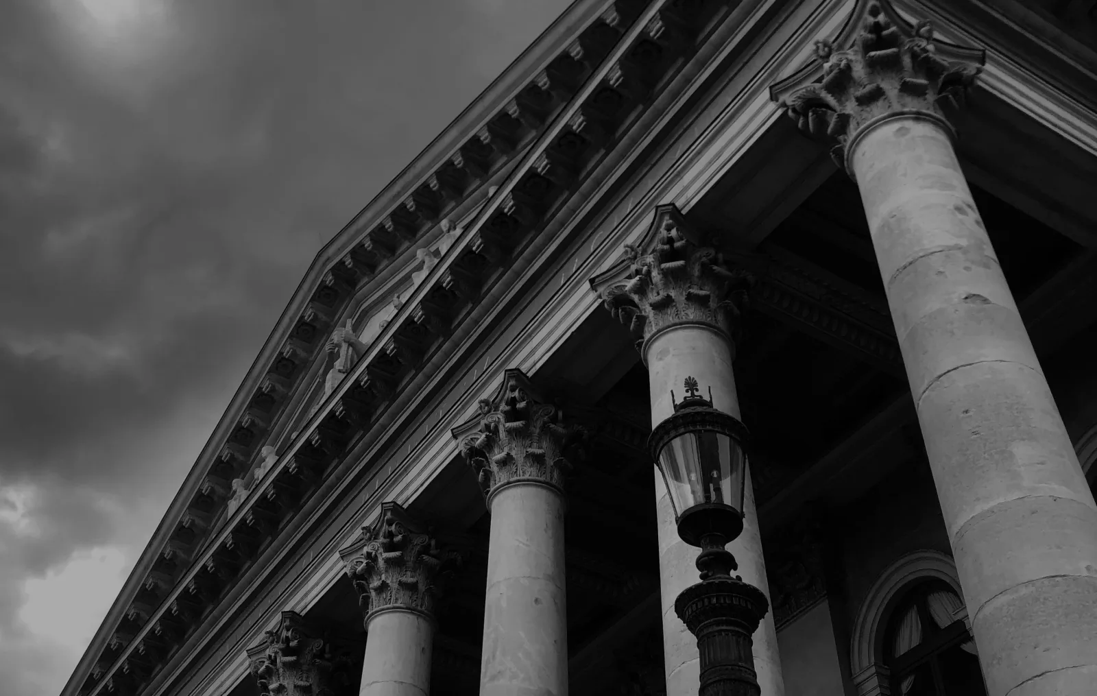 A low-angle view of a neoclassical courthouse pediment and Corinthian column against a stormy overcast sky - the weight of legal deliberation before public response