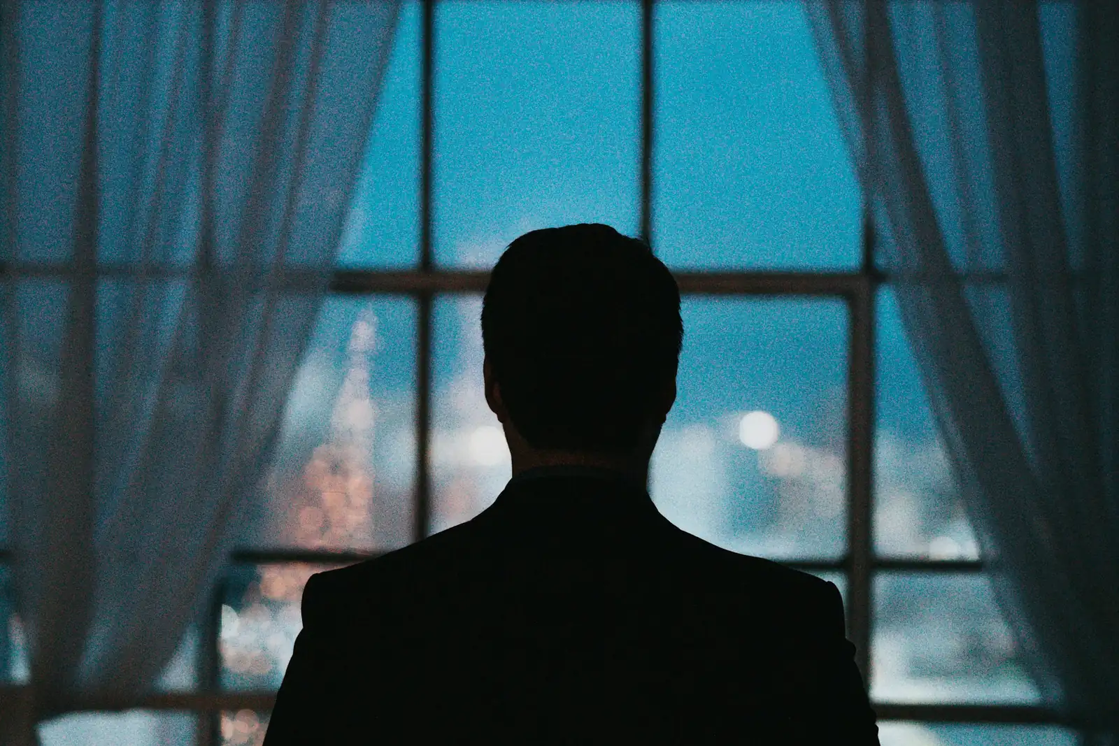 A suited figure silhouetted against a window at night, the city beyond in soft focus - an outline is all the room has before the person speaks