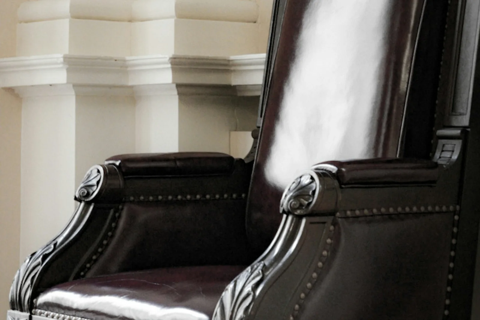 An empty high-back leather chair with carved arms and brass studding beside pale classical panelling - the executive seat, and the reputation no one is assigned to manage